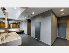 Moderner, gut beleuchteter Waschraum mit grauen Fliesen an Böden und Wänden, mehreren weißen Waschbecken mit Spiegeln darüber und Holzverkleidungen darunter. Auf der rechten Seite befinden sich flache, schwarze Heizkörper an den Wänden.