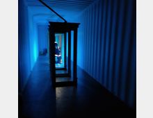Ein schmaler, länglicher Raum mit blauen Wänden und einer blauen Beleuchtung, vermutlich in einem Frachtcontainer. In der Mitte des Raumes befinden sich dunkle Rahmen, und im Hintergrund ist eine Person in schwachem Licht erkennbar.