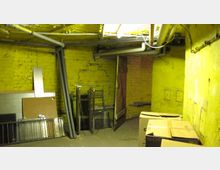 Ein Kellerraum mit gelb gestrichenen Ziegelwänden, sichtbaren Rohren unter der Decke und einer offenen Holztür. Im Raum stehen Kartons und Metallgegenstände an den Wänden lehnen.