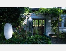 Eingang eines kleinen Hauses mit einer Pergola, die von Kletterpflanzen und grünem Blattwerk umgeben ist. Im Vordergrund befinden sich Gartenpflanzen, Laternen und Rosen, während im Hintergrund der Innenraum mit großen Fenstern sichtbar ist.