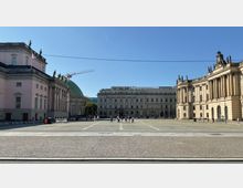 Weitläufiger, gepflasterter Platz mit umgebenden historischen Gebäuden. Links ein rosa Gebäude mit Säulen, rechts ein gelbliches Gebäude mit monumentalen Säulen, und zentral ein klassizistisches Gebäude; im Hintergrund eine grüne Kuppel und ein Baukran.