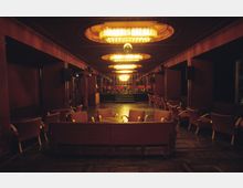 Ein gedämpft beleuchteter Ballsaal mit dunklem Holzboden, roten Wänden und zahlreichen Sitzecken mit gepolsterten Stühlen. Die Decke ist mit mehreren großen, beleuchteten Kronleuchtern verziert, und am hinteren Ende des Raumes befindet sich eine kleine Bühne mit roten Stühlen.