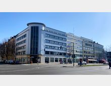 Eine Straßenecke mit einem mehrstöckigen Gebäude im Stil der modernen Architektur der Mitte des 20. Jahrhunderts, gekennzeichnet durch eine abgerundete Fassade mit vertikalen Fenstern. Im Erdgeschoss befinden sich Geschäfte und Büros, während die oberen Stockwerke überwiegend mit rechteckigen Fenstern ausgestattet sind.