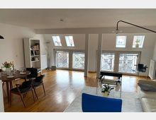 Helles Wohnzimmer mit Holzfußboden, Dachschrägen und mehreren Fenstern mit dekorativen Gitterelementen, die Tageslicht hineinlassen. Der Raum ist möbliert mit einem Esstisch, Stühlen, einem Sofa und modernen Sesseln; ein Bücherregal und Pflanzen ergänzen die Einrichtung.