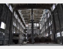 Ehemalige Industriehalle mit hohen Wänden aus dunklem Backstein und großen, vergitterten Fenstern. Im Inneren befinden sich rostige Tanks, Stahlträger und teilweise erhaltene Maschinenstrukturen, die eine verlassene Fabrikumgebung zeigen.