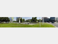 Grüne Parkanlage mit Rasenflächen, Bäumen und einem kleinen Brunnenplatz mit Sitzbänken im Vordergrund, umgeben von modernen Bürogebäuden. Im rechten Bildbereich verläuft eine mehrspurige Straße mit parkenden und fahrenden Autos und Fahnenmasten entlang des Gehwegs.