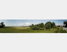 Weite Wiesenlandschaft mit vereinzelten Bäumen und kleinen Wasserflächen, im Hintergrund einige Gebäudestrukturen am Rand einer Straße. Der Himmel ist weitgehend klar mit wenig Wolken.