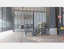 Industriehalle mit einem mehrstöckigen, verglasten Kontrollraum. Eine Metalltreppe führt zu einem erhöhten Bereich, und die Umgebung umfasst hohe Wände, große Fenster und gekennzeichnete Bodenmarkierungen.