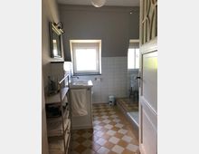 Kleines Badezimmer mit einem rechteckigen Fenster, einem Waschbecken samt Spiegel, sowie einer begehbaren Dusche mit Glastrennwand. Der Boden ist mit gelb-weißen Schachbrettfliesen bedeckt, und ein Regal mit Handtüchern steht links neben dem Waschbecken.