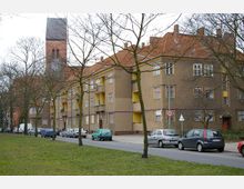 Straßenszene mit einer Reihe von mehrstöckigen Wohngebäuden in Braun und Gelb, einem roten Ziegeldach und Balkonen. Im Hintergrund ist ein Kirchturm aus Backstein sichtbar, während parkende Autos und Bäume die Straße säumen.