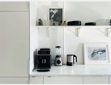 Minimalistische Küchenzeile mit weißer Arbeitsplatte, darauf eine schwarze Kaffeemaschine, ein silberfarbener Mixer und ein Wasserkocher. Oben ein weißes Regal mit schwarzen Tellern, Gläsern und Dekorationsobjekten.