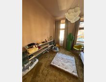 Ein schlicht eingerichtetes Zimmer mit beigefarbenen Wänden, einem Teppichboden und großen Fenstern mit Vorhängen. Im Raum stehen ein niedriges Regal mit Spielzeug und Textilien, eine grüne dekorative Kaktusfigur, zwei Stühle, sowie eine große, flache Matratze auf dem Boden.