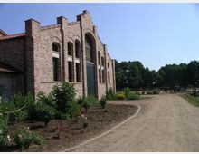Backsteinbau mit hohen Rundbogenfenstern und Satteldach, umgeben von gepflegten Beeten und einem unbefestigten Weg. Im Hintergrund stehen Bäume und eingezäunte Pferde auf einer Koppel.