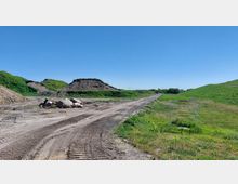 Eine offene Baustellenlandschaft mit grasbewachsenen Hügeln und einem unbefestigten Weg, der durch die Szene führt. Im Vordergrund befinden sich große Steine sowie Reifenspuren, und rechts ist eine Metallkonstruktion in einer Grünfläche sichtbar.
