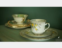 Zwei Porzellantassen mit Blumendekor stehen auf passenden Untertassen vor einer dunkelgrünen Wand. Die vordere Tasse zeigt gelbe Rosen, während die hintere mit goldenen Akzenten verziert ist.