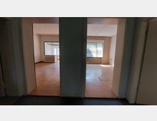 Blick in zwei leere, nebeneinanderliegende Zimmer mit Holzböden, großen Fenstern und Heizkörpern unter den Fenstern. Die Räume sind durch Türöffnungen von einem dunkleren Flur aus sichtbar.