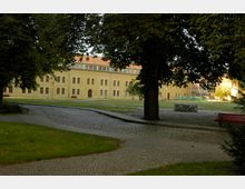 Innenhof mit gepflasterten Wegen, Rasenflächen und einem Baum im Vordergrund, im Hintergrund ein großes, gelbliches historisches Backsteingebäude mit vielen Fenstern und einem roten Ziegeldach.