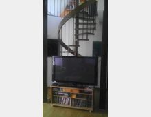 Wohnzimmer mit hölzernem Fernsehschrank, darauf ein Flachbildfernseher. Im Hintergrund befindet sich eine Wendeltreppe aus Holz und Metall, die zu einem oberen Bereich führt.