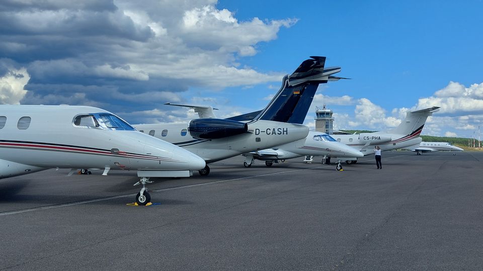Mehrere Geschäftsflugzeuge stehen nebeneinander auf dem Vorfeld eines Flughafens, mit sichtbaren Fahrwerkskeilen auf dem Asphalt. Im Hintergrund sind ein kleiner Kontrollturm sowie eine flache, grüne Umgebung unter einem wolkigen Himmel zu sehen.