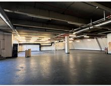 Ein leerer Industriebereich mit grauem Fußboden, sichtbaren Lüftungsrohren an der Decke und weißen Wänden. Der Raum ist spärlich beleuchtet durch Reihen von Deckenleuchten, und ein einzelner Holzrollwagen steht in der Mitte.