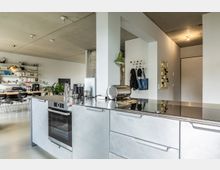 Offene, moderne Küche mit einer Arbeitsinsel aus Edelstahl, integriertem Backofen und Kochfeld. Im Hintergrund ein Essbereich mit Bücherregalen an der Wand, einer hölzernen Tafel und schwarzen Stühlen.