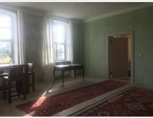 Heller Raum mit grünen Wänden, großen Fenstern mit weißen Vorhängen und einem Holzboden, dekoriert mit orientalischen Teppichen. Links steht ein kleiner Holztisch mit Stühlen, in der Mitte ein filigraner Schreibtisch und rechts führt eine offene Tür in einen weiteren Raum.