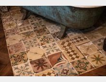 Ein Badezimmerboden mit mehrfarbigen ornamental bedruckten Fliesen in verschiedenen Mustern und Farben. Im Hintergrund ist der untere Teil einer altertümlichen, metallisch-grünen Badewanne mit Beinen zu sehen.