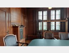 Holzvertäfelter Innenraum eines historischen Besprechungszimmers mit großen, bleiverglasten Fenstern. Zu sehen sind ein Kamin, ein hölzernes Wandpaneel mit Namensgravuren, Ledersessel und ein Tisch im Vordergrund.