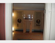 Ausstellungsraum mit Holzfußboden, einem beleuchteten Modell auf einem hölzernen Podest und Schautafeln an den Wänden mit Bildern und Texten, die historische Informationen zeigen.