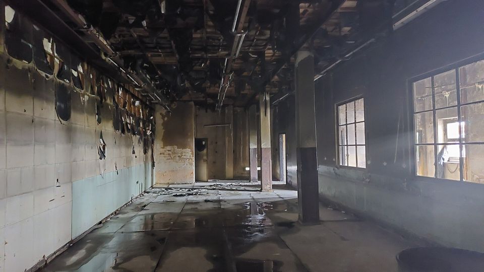 Ein verlassener Raum mit beschädigten Wänden und Decken, teilweise mit verbranntem Material. Die Fensterscheiben an der rechten Seite lassen Licht in den ansonsten dunklen Raum fallen, und der Boden scheint feucht oder nass zu sein.