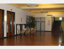 Ein moderner Innenbereich mit glänzendem roten Marmorboden und gelben Decken mit runden, hängenden Leuchten. Links befinden sich große Holztüren, rechts steht eine Sitzgruppe mit Korbstühlen und einem kleinen Tisch vor einem großen Fenster, im Hintergrund führt eine Treppe nach oben.