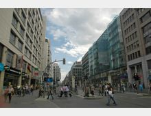 Straßenszene in einer belebten Stadt mit modernen Geschäftsgebäuden aus Glas und Stein auf beiden Seiten. Fußgänger, Radfahrer und Ampeln prägen das Bild, während am linken Gebäude mehrere Flaggen hängen.