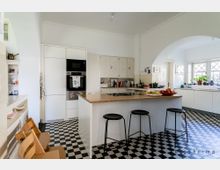 Geräumige Küche mit einem zentralen Küchenblock mit Holzarbeitsplatte und drei schwarzen Hockern. Weiß gestrichene Wände, Schachbrettmuster-Fliesen am Boden, eingebaute Küchenschränke und große Fenster mit Sprossen sorgen für viel Licht.