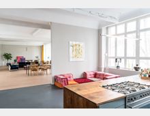 Offener Wohnraum mit moderner Einrichtung, bestehend aus einer Küche mit Gasherd und Holzarbeitsplatte im Vordergrund, einer gemütlichen Sitzecke mit bunten Sofas rechts sowie einem Ess- und Wohnbereich mit Holzmöbeln und großen Fenstern im Hintergrund. Helle Wände und viel Tageslicht sorgen für eine klare, freundliche Atmosphäre.