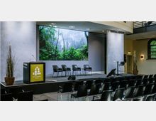 Ein moderner Veranstaltungsraum mit einer Bühne, auf der mehrere schwarze Stühle und ein Rednerpult mit dem Logo „Besondere Orte“ stehen. Im Hintergrund befindet sich eine große Leinwand mit einem Bild eines dichten Waldes, während vor der Bühne mehrere Reihen schwarzer Stühle für das Publikum angeordnet sind.