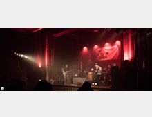 Eine Live-Bühne in einem dunklen Veranstaltungsraum, beleuchtet mit rotem und warmem Licht. Eine Band tritt auf, umgeben von Instrumenten, Verstärkern und einem Banner mit dem Schriftzug „The Dirty Franks“ im Hintergrund.