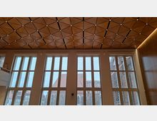 Innenraum eines modernen Saals mit hoher Decke aus dreieckigen Holzpaneelen und punktförmigen Leuchten. Große, hohe Fensterfronten mit gemustertem Glas lassen Tageslicht herein und geben den Blick auf Backsteinbauten und Kirchturmstrukturen draußen frei.