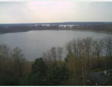 Blick auf einen See, umgeben von kahlen Bäumen und dichter Vegetation am Ufer. Im Vordergrund sind Bäume und ein teilweises Dach eines Gebäudes sichtbar, während im Hintergrund der See in eine flache, bewaldete Landschaft übergeht.