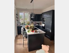 Moderne Küche mit dunkelgrauen Schränken, weißer Arbeitsplatte und zentraler Kochinsel. Ein großes Fenster lässt Tageslicht herein, und die Einrichtung umfasst hängende Glaslampen sowie dekorative Elemente wie Blumen und Obstschalen.