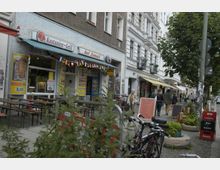 Straßenszene mit einem Imbiss namens „Kastanien-Grill bei Ismail“ in einem Altbaugebäude mit grauer Fassade. Vor dem Imbiss stehen Holzbänke, daneben Fahrräder und Pflanzen; Passanten schlendern an weiteren Geschäften und Cafés entlang, während ein Schild mit einer Speisekarte am Gehweg steht.
