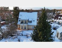 Luftaufnahme eines hellen, zweigeschossigen Wohnhauses mit steilem, schneebedecktem Satteldach und mehreren Gauben. Das Haus liegt in einem verschneiten Garten mit Zaun, umgeben von hohen Nadel- und Laubbäumen sowie benachbarten Gebäuden.