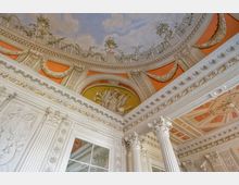 Dekorierter Innenraum mit barocker Architektur; reich verzierte Säulen mit korinthischen Kapitellen, Stuckornamente und ein bemalter Deckenhimmel mit Wolken und Pflanzenmotiven. Elemente in Weiß, Orange und Gold prägen das Erscheinungsbild.
