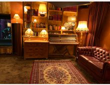 Gemütlicher Innenraum im Retro-Stil mit einem roten Chesterfield-Sofa, einem offenen Klavier, mehreren antiken Tischlampen, hölzernen Bücherregalen und dunklen Vorhängen. Ein verziertes Teppichmuster bedeckt den Fußboden.