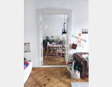 A cozy living and dining space with light-colored walls, wooden floors, and an arched door frame separating the rooms. The dining area features a wooden table and chairs with potted plants and framed art in the background, while the living space includes a leather sofa and scattered toys and decorations.