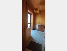 Holzverkleideter Flur mit hellem Holzboden, einem kleinen Schrank und einer Kommode. An der Decke hängt eine gelbe Lampe, und im Hintergrund befinden sich ein Heizkörper, ein Metallschuhregal und eine Tür mit Glaseinsatz, die nach außen führt.