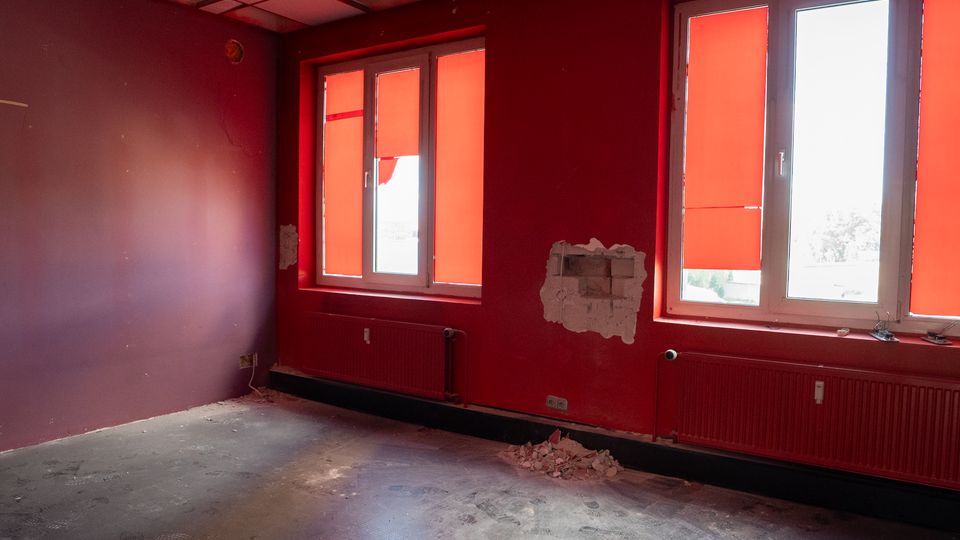 Leerer Innenraum mit dunkelroten Wänden und zwei großen Fenstern, die mit roten Rollos abgedeckt sind; unter den Fenstern verlaufen Heizkörper. An der Wand sind beschädigte Putzstellen und Bauschutt am Boden sichtbar, die Decke besteht aus Deckenplatten.