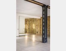 Innenraum im Loft-Stil mit freiliegender Backsteinwand, poliertem Betonboden und großer spiegelnder Schrank- bzw. Wandfläche aus Glaspaneelen. Rechts steht eine massive, genietete Stahlstütze, ergänzt durch schlichte Wandbeleuchtung und eine dunkle Tür.