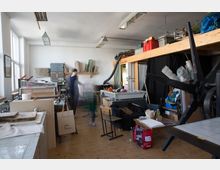 Ein heller Künstler- oder Druckereiraum mit Holzfußboden, großen Fenstern und zahlreichen Werkmaterialien wie Rahmen, Papierstapeln und einer großen Druckmaschine im Vordergrund; im Hintergrund befinden sich Regale mit weiteren Utensilien und eine erhöhte Holzplattform.