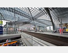 Hauptbahnhof mit modernen Glas- und Stahlbögen, überdachenden Dächern und mehreren Bahnsteigen. Im Vordergrund sind ein gläserner Fahrstuhl, Zuganzeigen und Rolltreppen sichtbar, während Passagiere auf den Bahnhöfen warten.