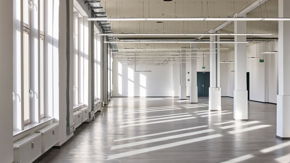 Großer, leerer Innenraum mit hohen Decken, weißen Wänden und vielen bodentiefen Fenstern auf der linken Seite, die viel Tageslicht hereinlassen. Die Decke verfügt über sichtbare Kabeltrassen und schmale, rechteckige Leuchten, während der Boden aus glattem Beton besteht.